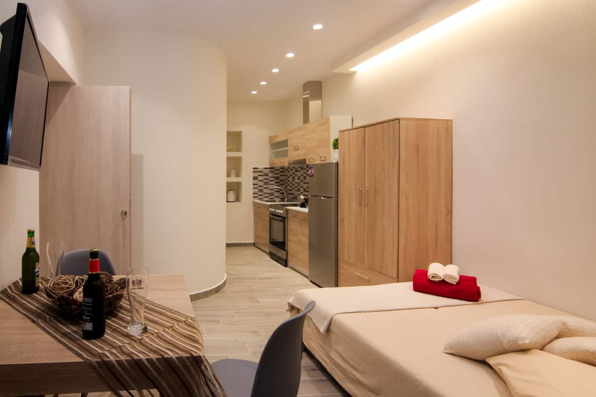 A modern hotel room with a bed, a small desk with a bottle and glass, a wooden wardrobe, and a kitchenette in the background.