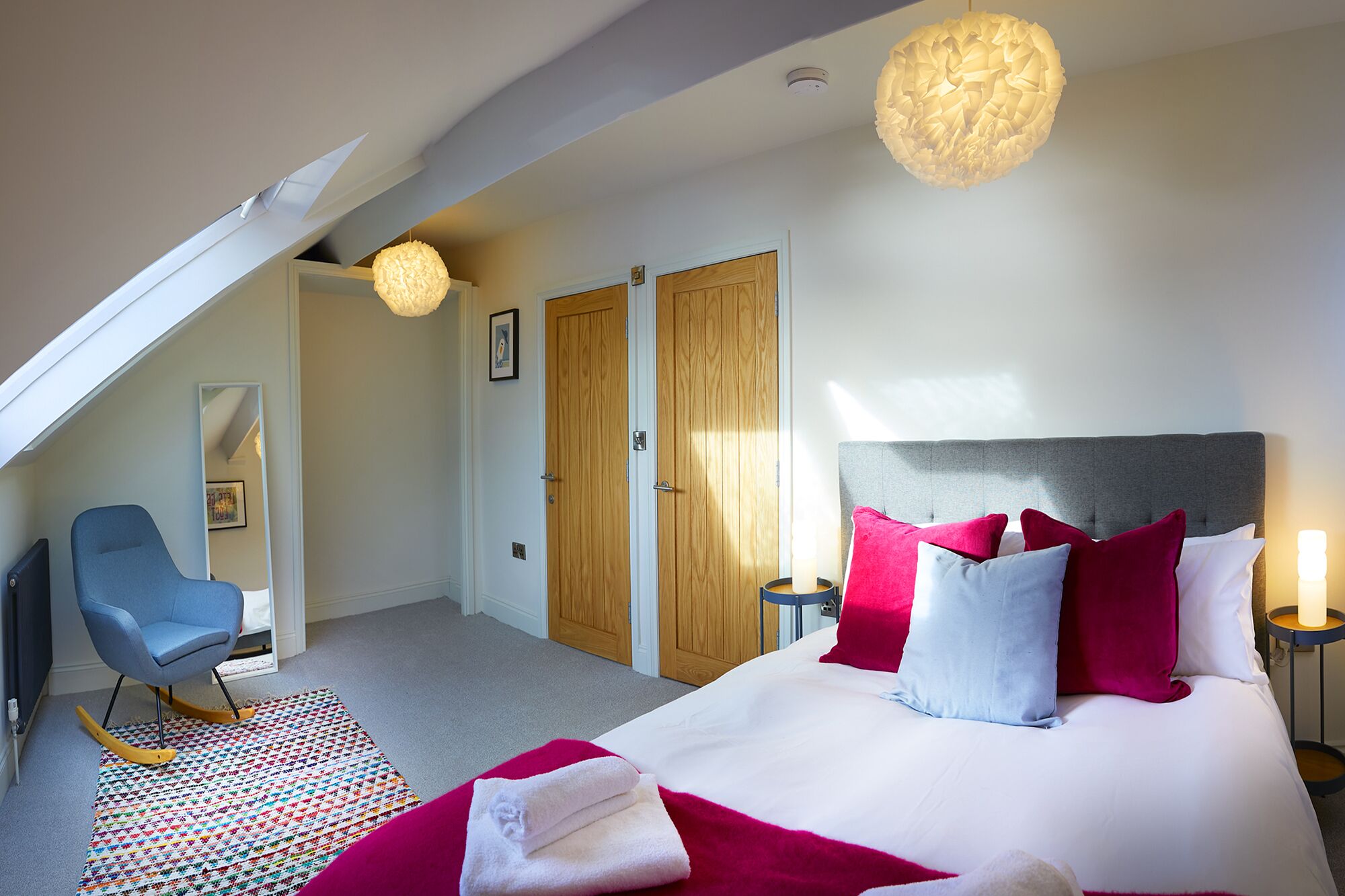 Une chambre à coucher avec un grand lit, un fauteuil à bascule, un tapis coloré et un plafond en pente doté d’une lucarne.