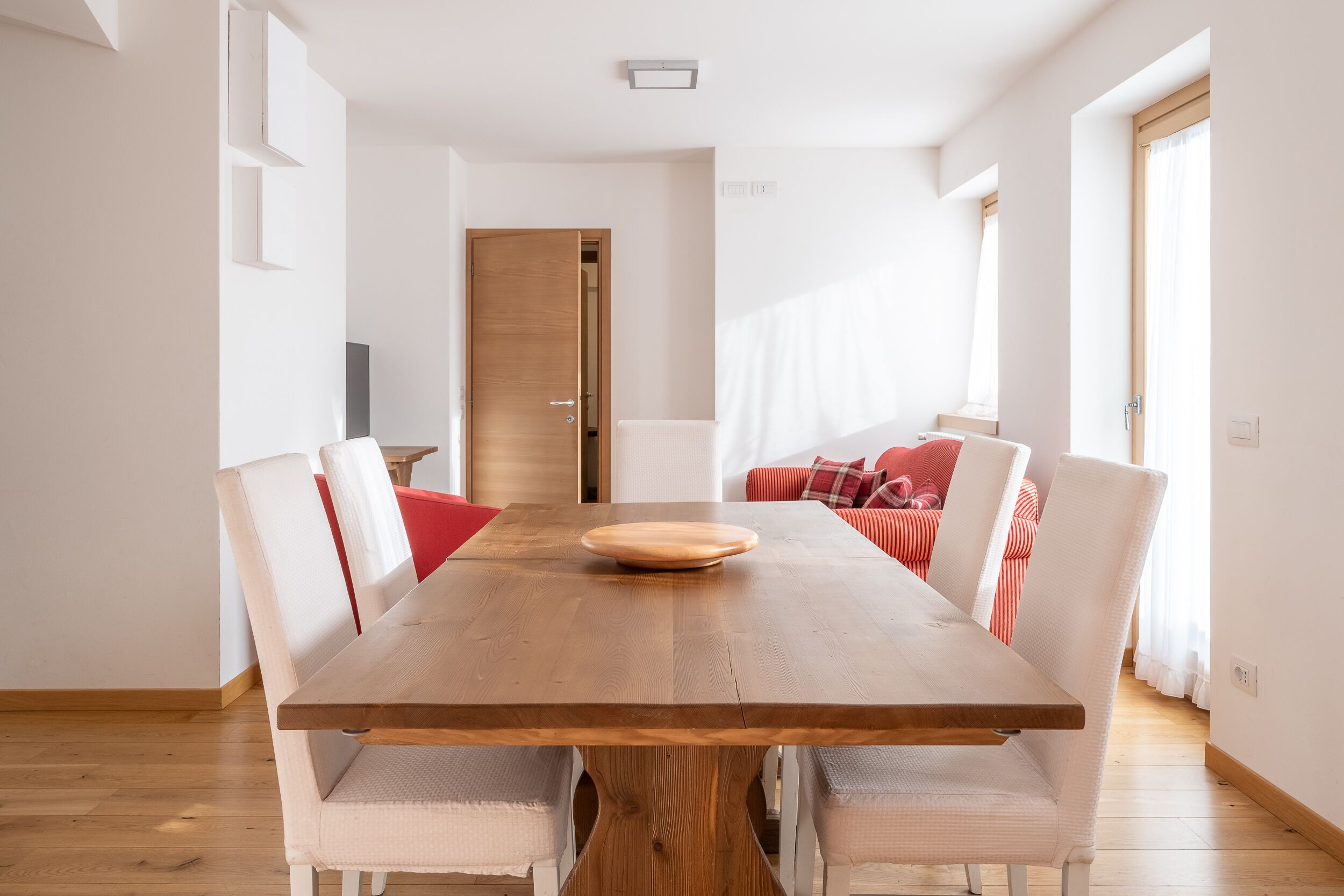 Zona pranzo con tavolo in legno, sedie bianche e divano rosso.