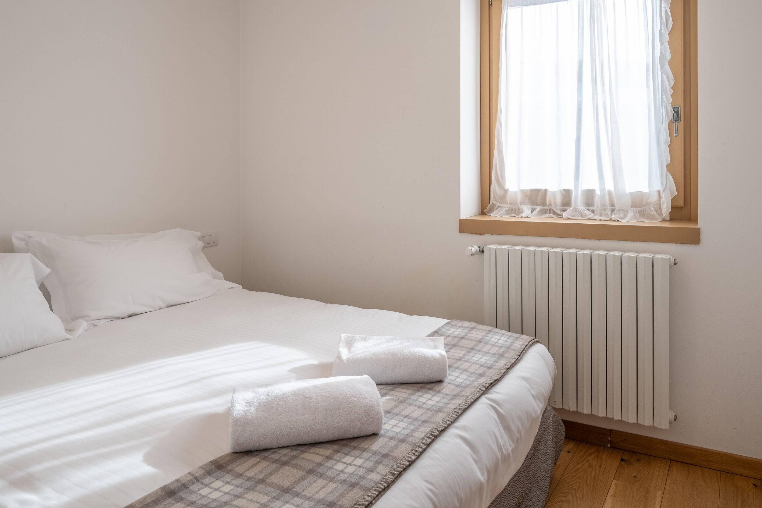 Una cama bien hecha con sábanas blancas y una manta escocesa, una ventana con cortinas traslúcidas y un radiador blanco.