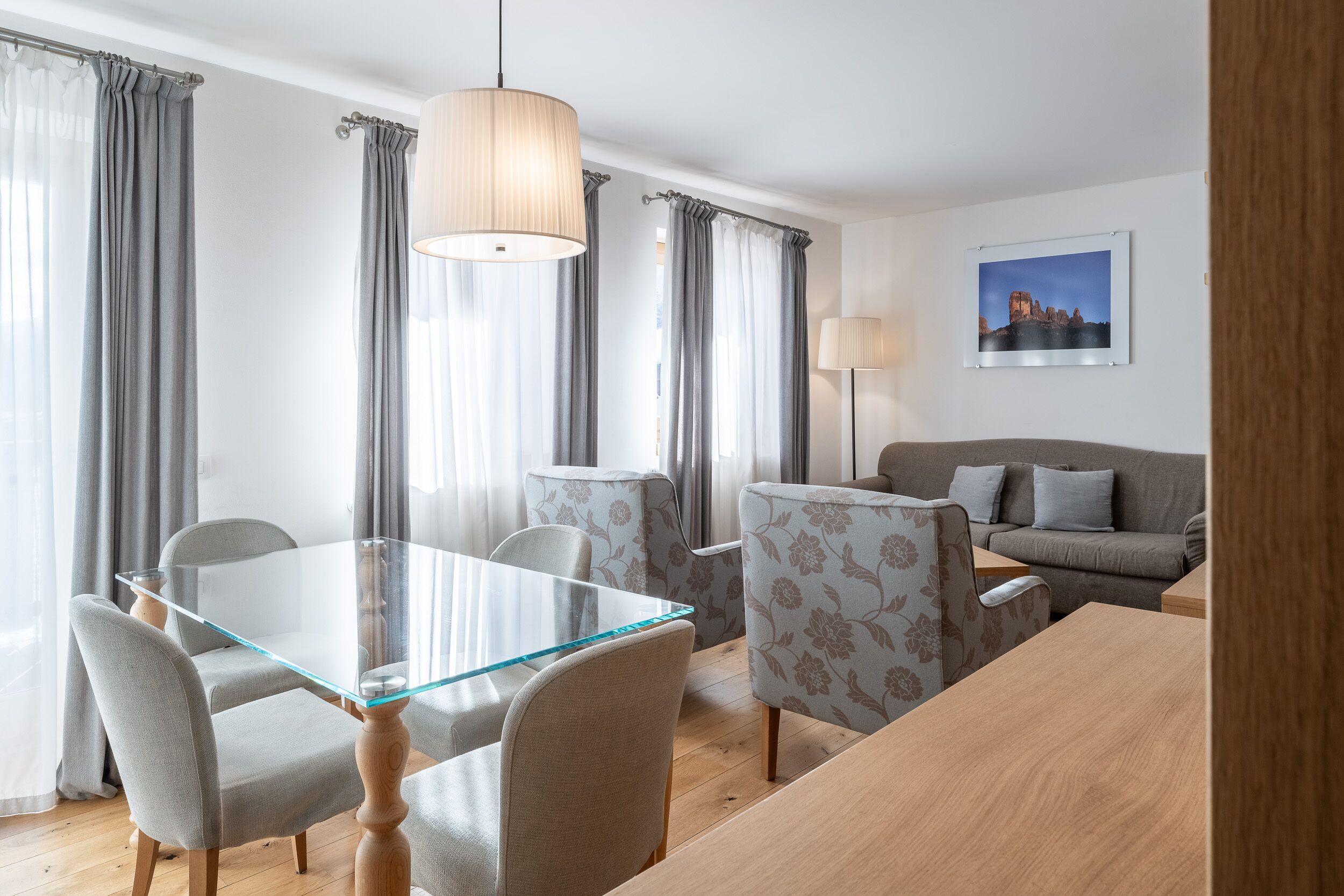 A modern living room with a glass dining table, grey upholstered chairs, a grey sofa, and a large window with grey curtains.