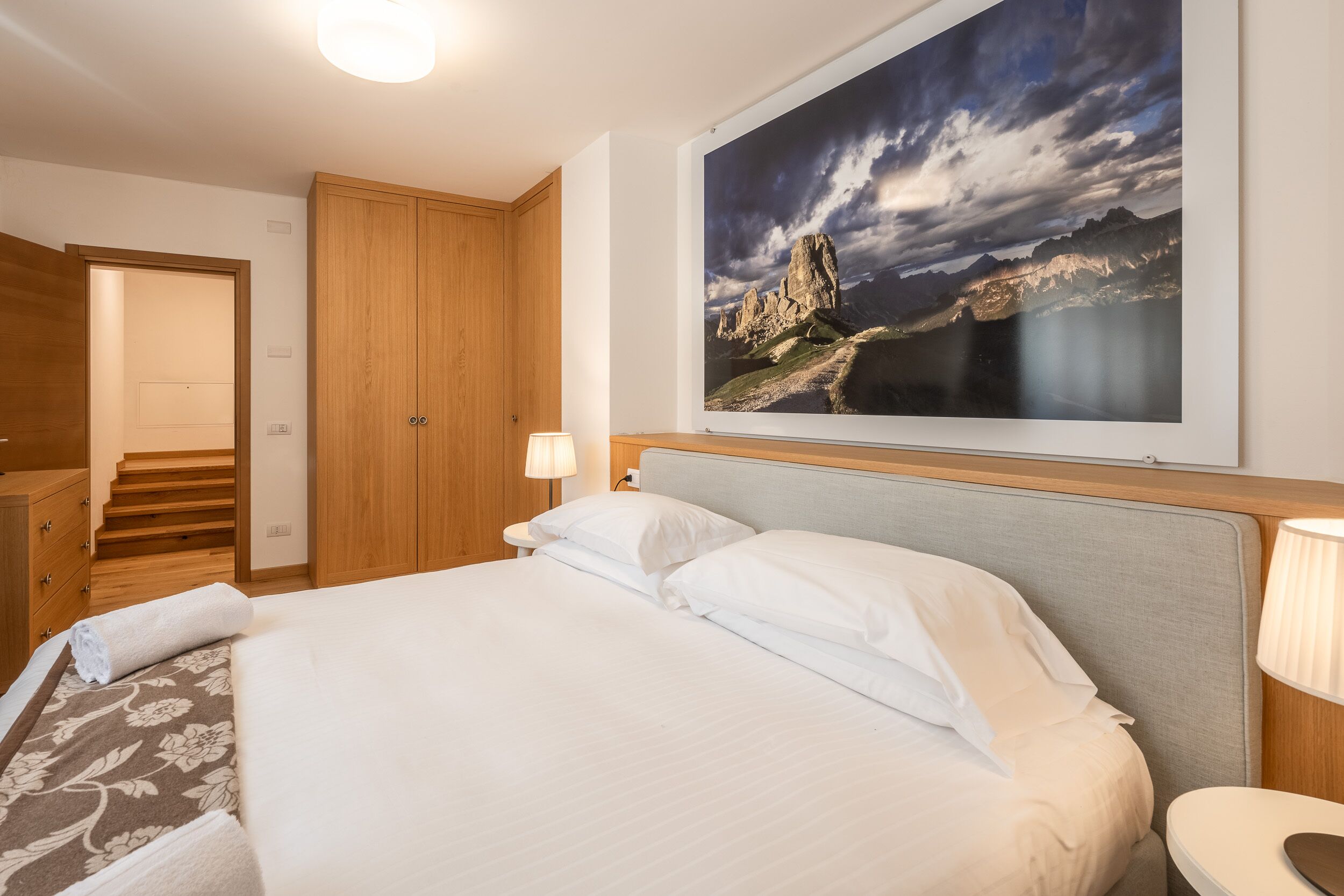 A hotel room with a bed, a nightstand, a lamp, a wooden wardrobe, and a framed landscape painting.