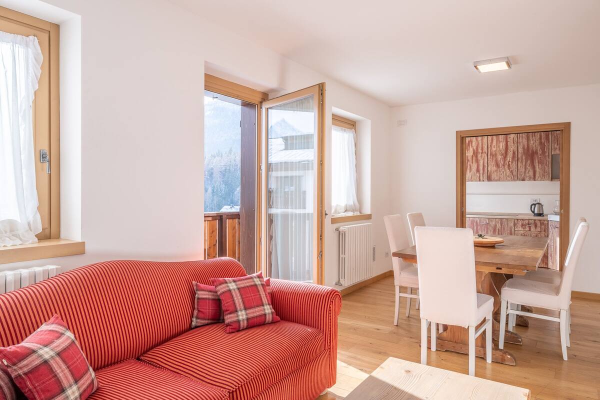 A cozy living room with a red-striped sofa, a dining table with white chairs, and a view of a mountain through a large window.