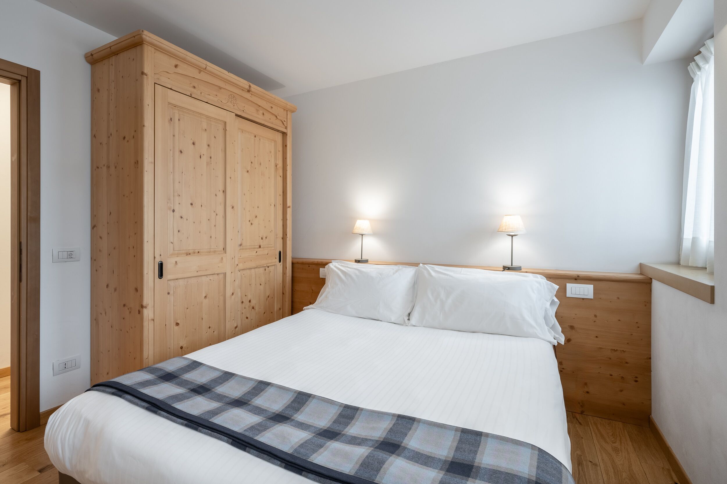 Ein Schlafzimmer mit großem Holzschrank, zwei Nachttischlampen und einer karierten Decke auf dem Bett.