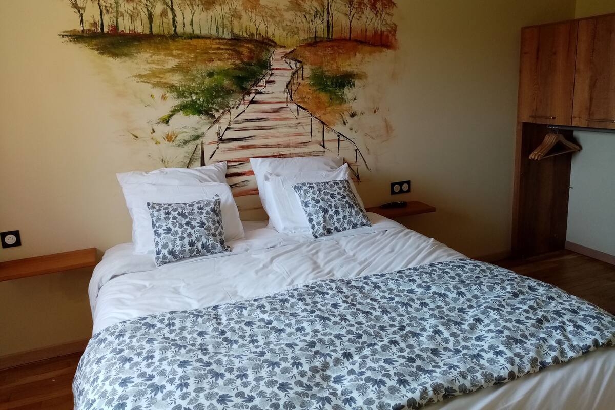 A bedroom with a bed, pillows, a nightstand, a wooden wardrobe, and a wall mural.