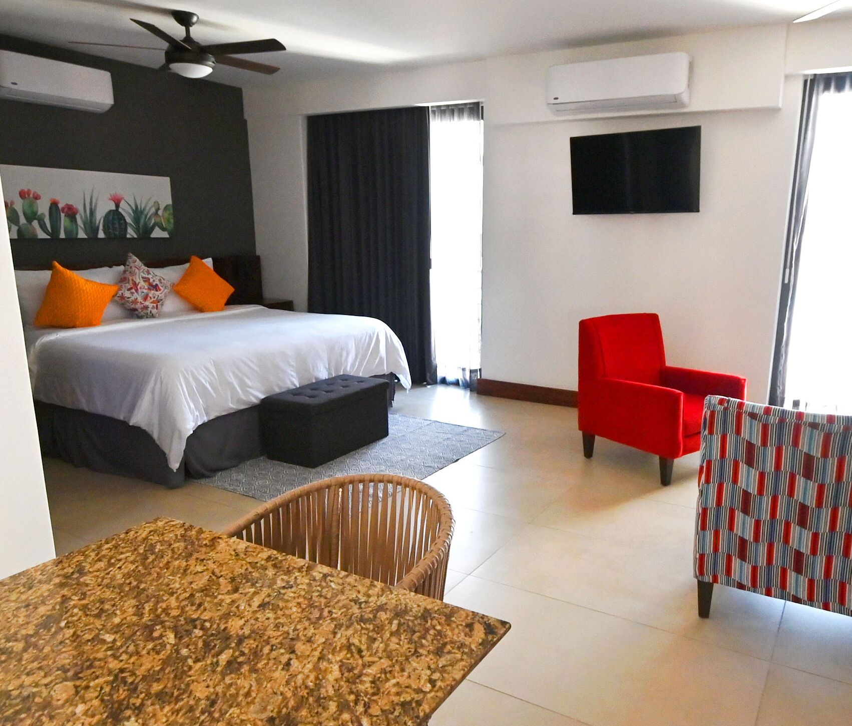 A hotel room with a bed, a red armchair, a patterned table, a wicker chair, and a painting of cacti.