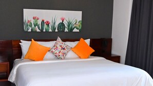 Une chambre d’hôtel avec un grand lit, du linge de lit blanc, deux oreillers orange, un oreiller à motifs floraux, une tête de lit sombre et un climatiseur fixé au mur.