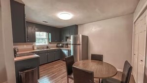 A compact kitchen with dark cabinets, a stainless steel refrigerator, and a round wooden dining table with chairs. - Cozy Cedartown Retreat
 (Cedartown)