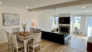 A modern living room with a dining table, chairs, a black sofa, a TV, and a fireplace. - Beachfront Paradise 10 steps from the Beach. Heated Pool, Spa, Sauna. No fees (Port Hueneme)