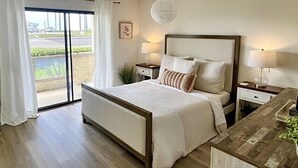 A modern bedroom with a large bed, two bedside tables with lamps, a view of the outdoors through a sliding door, and a decorative wall piece. - Beachfront Paradise 10 steps from the Beach. Heated Pool, Spa, Sauna. No fees (Port Hueneme)