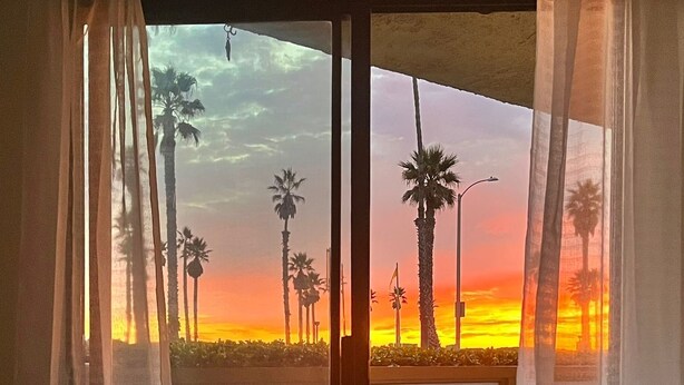 A room with a large sliding glass door leading to an outdoor balcony with a view of palm trees and a sunset. - Beachfront Paradise 10 steps from the Beach. Heated Pool, Spa, Sauna. No fees (Port Hueneme)