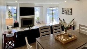 A modern living room with a fireplace, a flat-screen TV, and a dining area with a wooden table and chairs. - Beachfront Paradise 10 steps from the Beach. Heated Pool, Spa, Sauna. No fees (Port Hueneme)