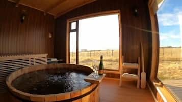 Un spa con paredes de madera, una bañera de hidromasaje, una mesa con una botella y vasos, y vistas al campo.