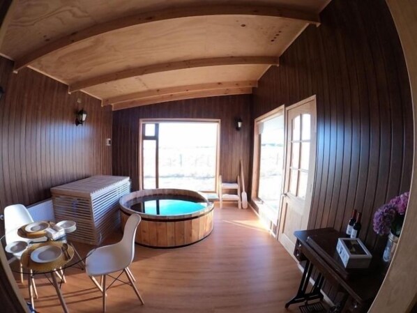 A wooden interior with a hot tub, a dining area with chairs and a table, and a bar area with bottles and glasses.