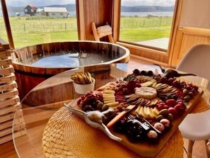 A table with a charcuterie board, a hot tub, and a view of a house and landscape. - Cabañas Las Lengas (Natales)