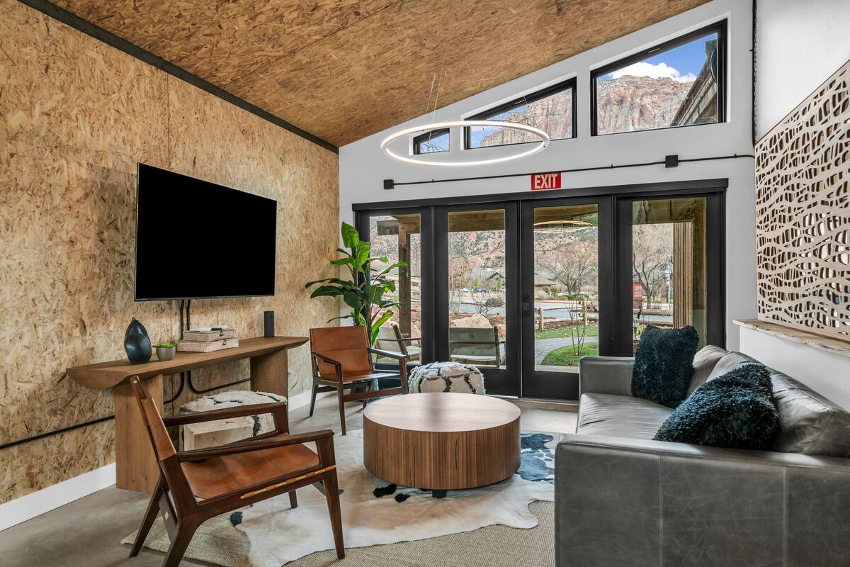 A modern interior with a wooden ceiling, a flat-screen TV, a round wooden coffee table, a sofa with blue cushions, and a view of the outdoors through large windows.