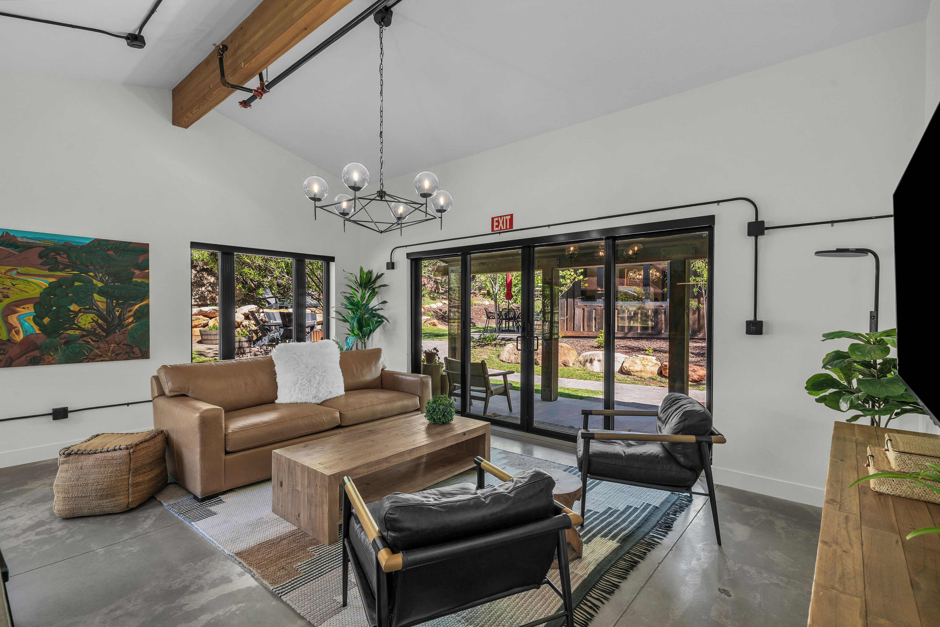 A modern living room with a brown leather sofa, a wooden coffee table, and a black chair. There is a large window with a view of an outdoor garden.