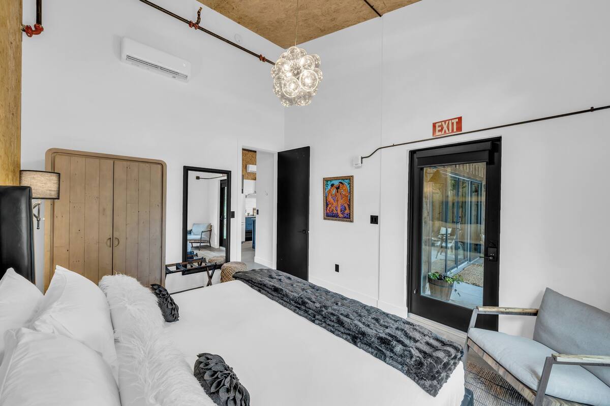 A modern hotel room with a large bed, a wooden wardrobe, a mirror, a black door, and an exit sign.