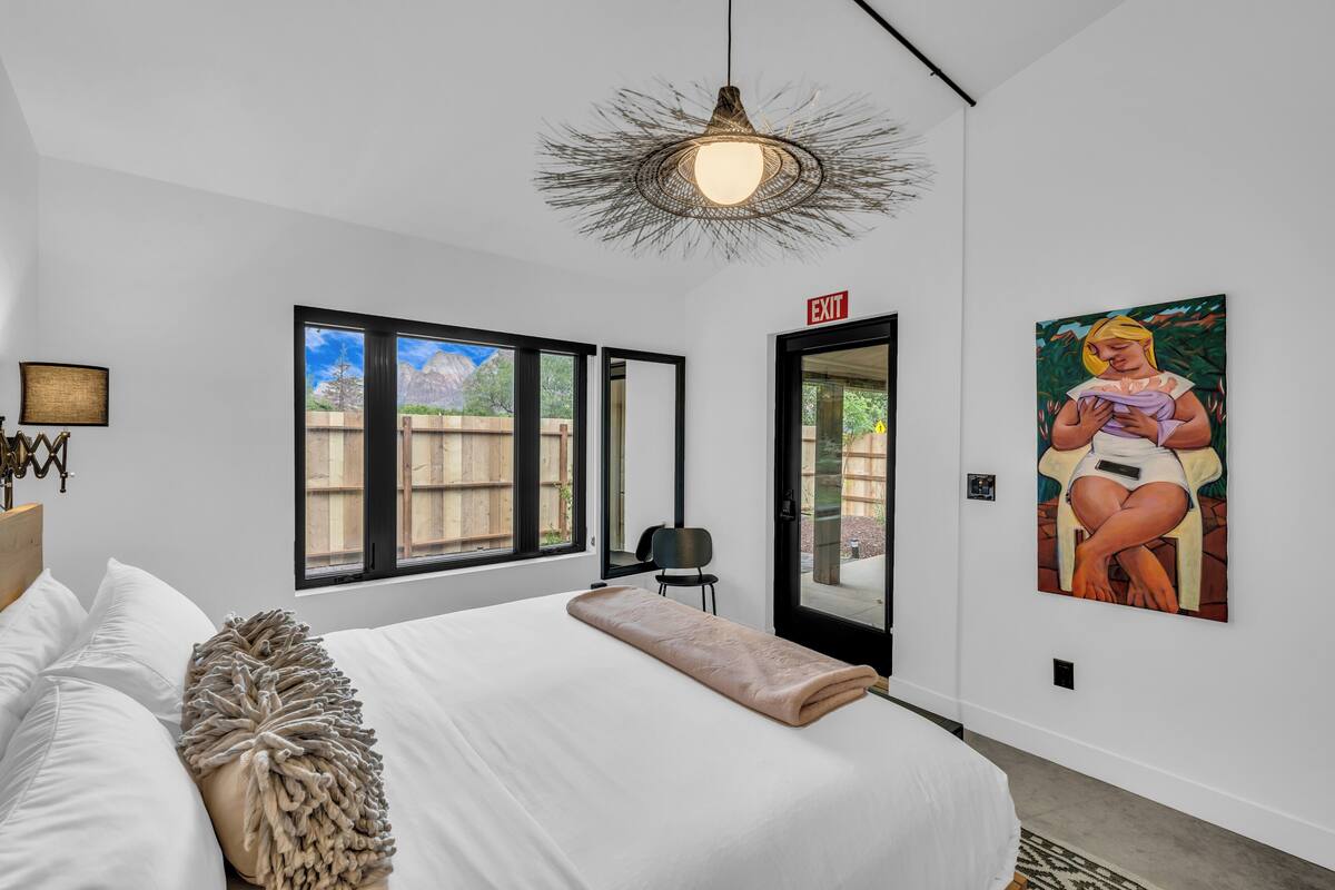 A modern bedroom with a large bed, a hanging light fixture, a painting on the wall, and a view of the outdoors through the window.