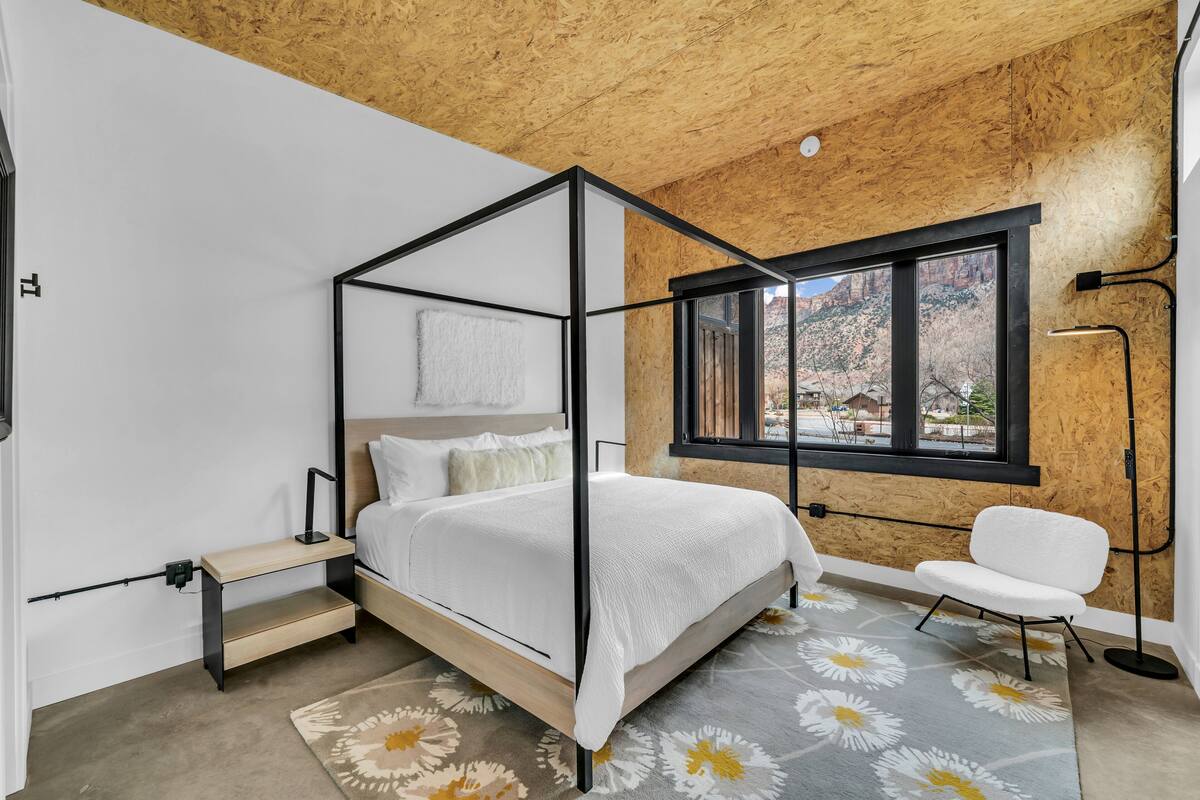 A modern bedroom with a four-poster bed, a wooden ceiling, a large window, and a white chair.