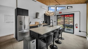 Una cocina moderna con un refrigerador de acero inoxidable, un horno empotrado y una isla grande con taburetes de bar. Se ve una ventana con vistas al exterior, y un letrero de salida sobre la puerta.