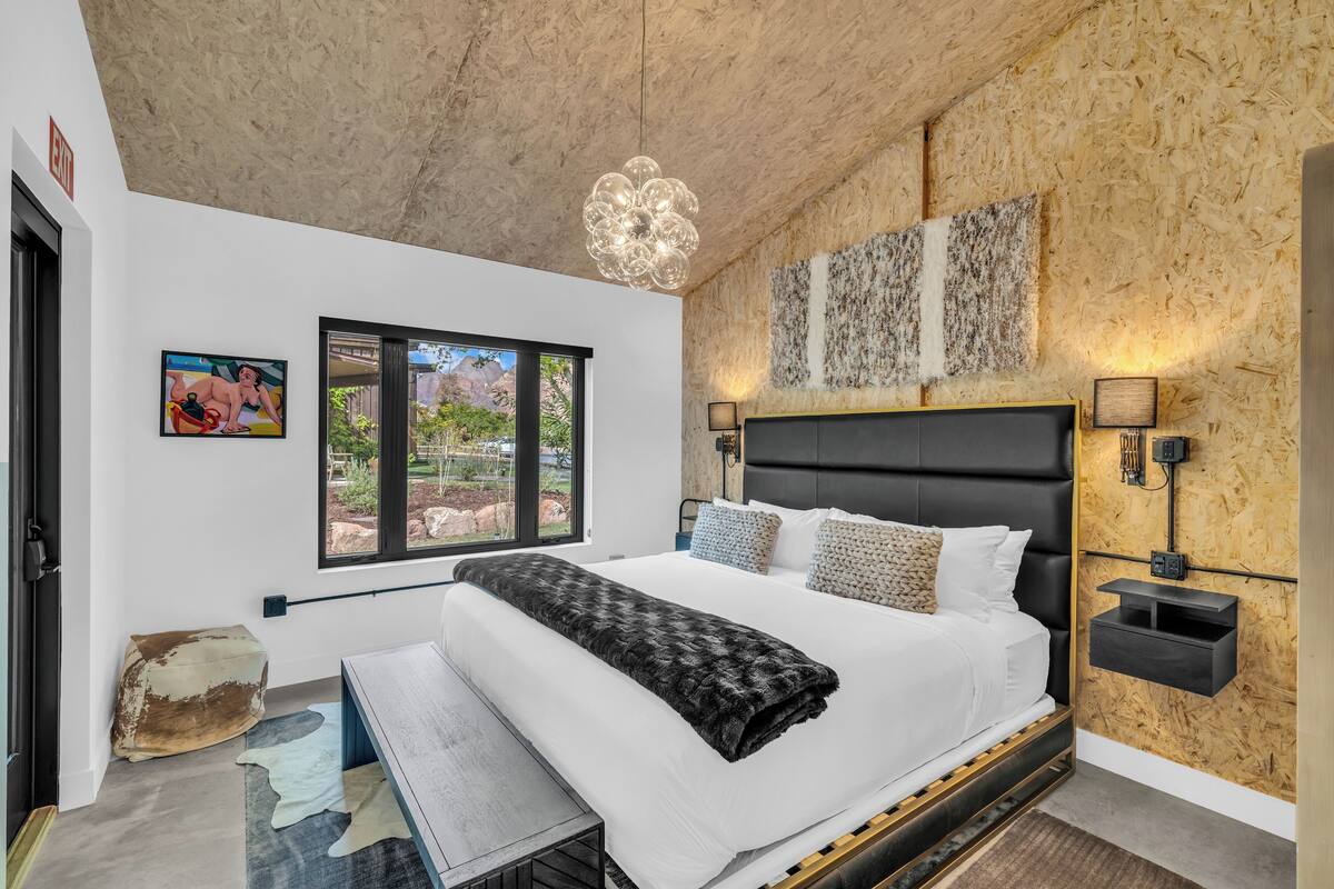 A modern bedroom with a large bed, a wooden ceiling, a window with a view, and a wall-mounted lamp.