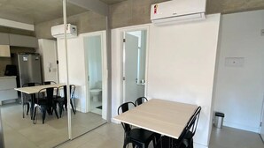 A modern kitchen with a refrigerator, a dining table with chairs, and a large mirror reflecting the room. - Novo Studio Prox. Av Paulista, Garagem e Academia (Consolação São Paulo)