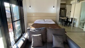 A modern bedroom with a large bed, a gray sofa with cushions, a ceiling light fixture, and a view of the outside through a large window. - Novo Studio Prox. Av Paulista, Garagem e Academia (Consolação São Paulo)