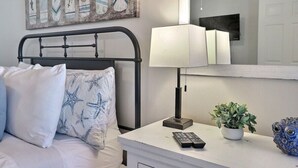 A bedroom with a bed, a bedside table with a lamp, a potted plant, and a decorative item. - 2BR Condo | mins to Beach & Sports Complex (Gulf Shores)