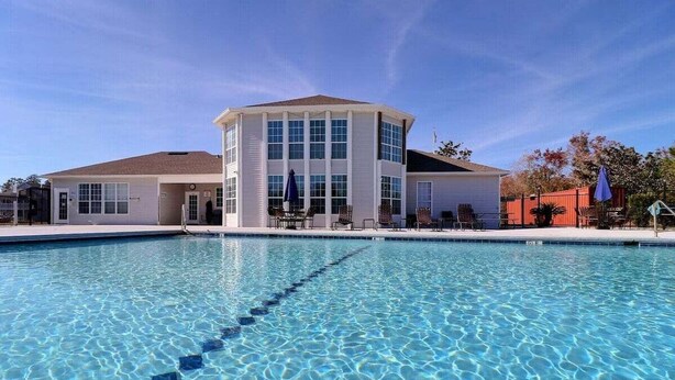 A swimming pool with a clear blue water surface, surrounded by a building with multiple windows and a covered seating area. - 2BR Condo | mins to Beach & Sports Complex (Gulf Shores)