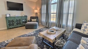 A living room with a flat-screen TV, a green cabinet, a grey armchair, a sofa, a coffee table, and a window with curtains. - 2BR Condo | mins to Beach & Sports Complex (Gulf Shores)