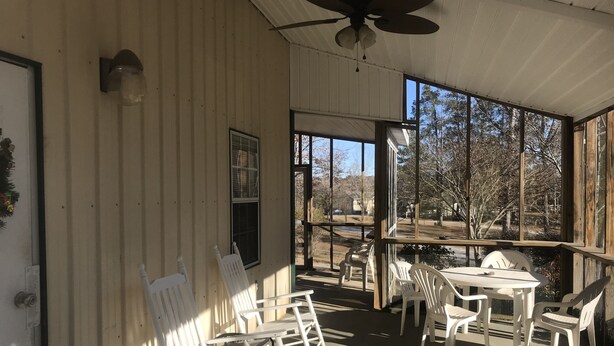 Un espace extérieur abrité avec des chaises et une table en plastique blanc, de grandes fenêtres offrant une vue sur l’extérieur et un ventilateur de plafond.