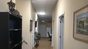 Un couloir avec une étagère noire, une plante en pot, un tableau encadré au mur et une porte donnant sur une pièce où se trouvent un bureau et une chaise.