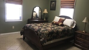 Une chambre à coucher comprenant un lit, une commode, une table de chevet, un miroir et deux fenêtres avec des rideaux.