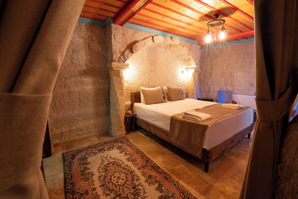 A bedroom with a stone wall, wooden ceiling beams, a bed with a wooden headboard, a nightstand, and a patterned rug.
