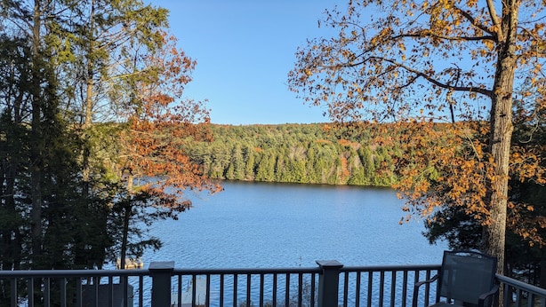 A deck with a table and chairs overlooking a lake surrounded by trees with autumn foliage. - Private Lakehouse Getaway for relaxing and fun! (Weare)