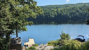 A lakeside cabin with a deck, surrounded by lush greenery and a forested hill in the background. - Private Lakehouse Getaway for relaxing and fun! (Weare)