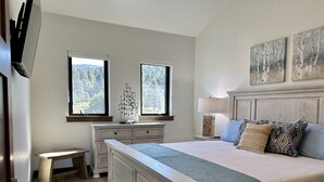 A bedroom with a large bed, a nightstand, a dresser, two windows with a view of trees, and a painting above the bed. - NEW 4BD, 4BA, Sleeps 13, Close to Ski Lifts, Sauna, Mtn Views (Angel Fire)