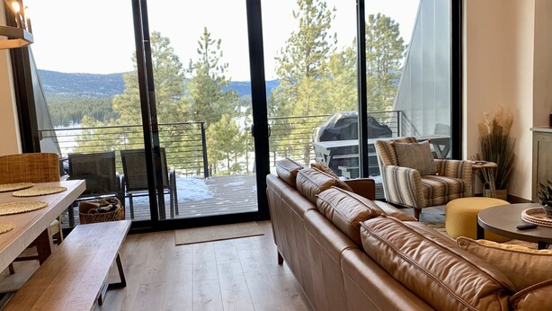 A modern living room with a large sliding glass door leading to a balcony, a leather sofa, a wooden dining table, and a small round table with a remote control. - NEW 4BD, 4BA, Sleeps 13, Close to Ski Lifts, Sauna, Mtn Views (Angel Fire)
