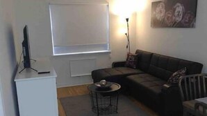 Un salon avec un meuble TV blanc, un canapé noir, une petite table d’appoint, un lampadaire et un tableau accroché au mur.