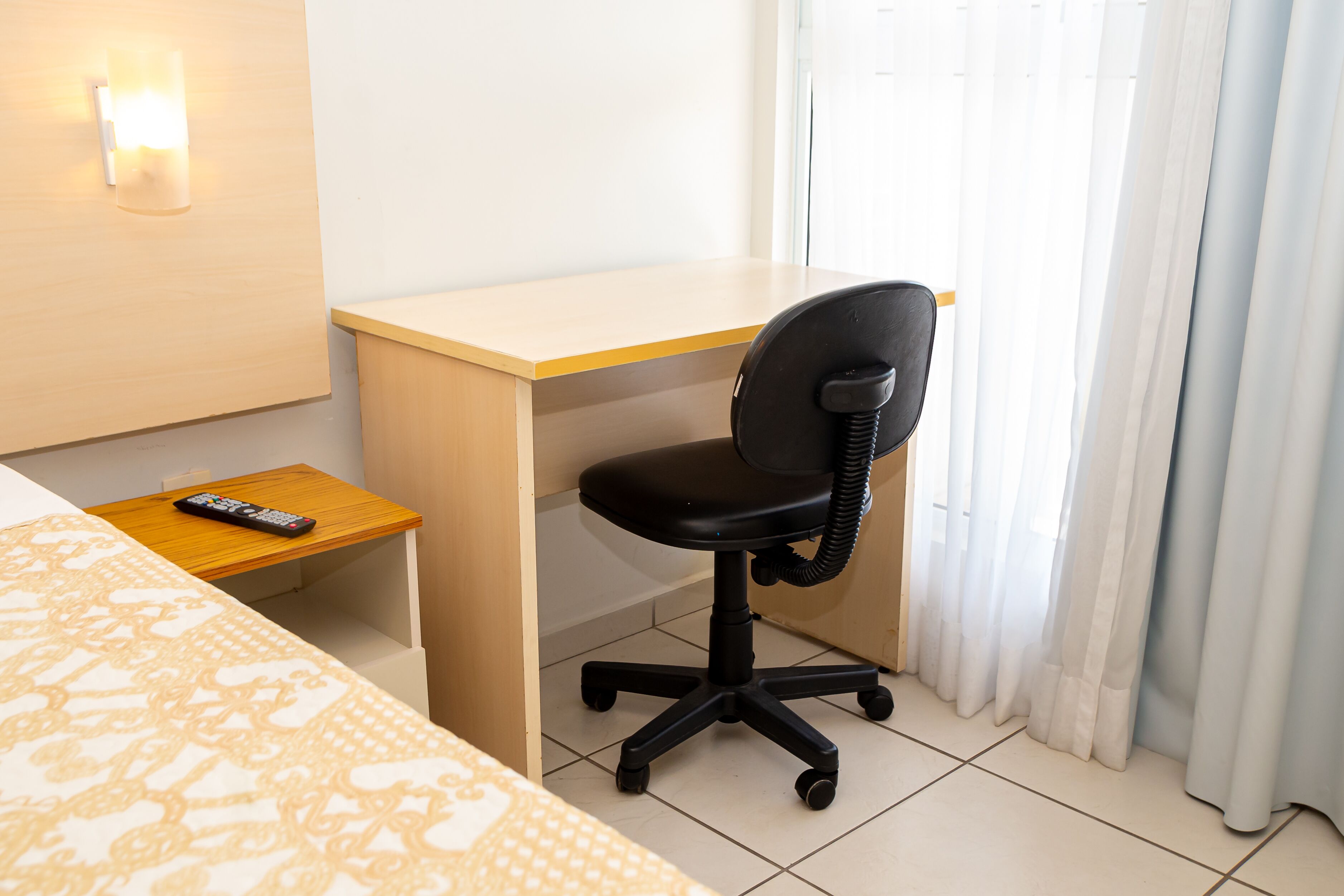 Een hotelkamer met een bed, een bureau, een stoel, een afstandsbediening en een raam met gordijnen.