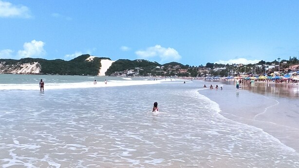 Una playa con gente en el agua y en la orilla, cielo despejado y colinas a lo lejos.