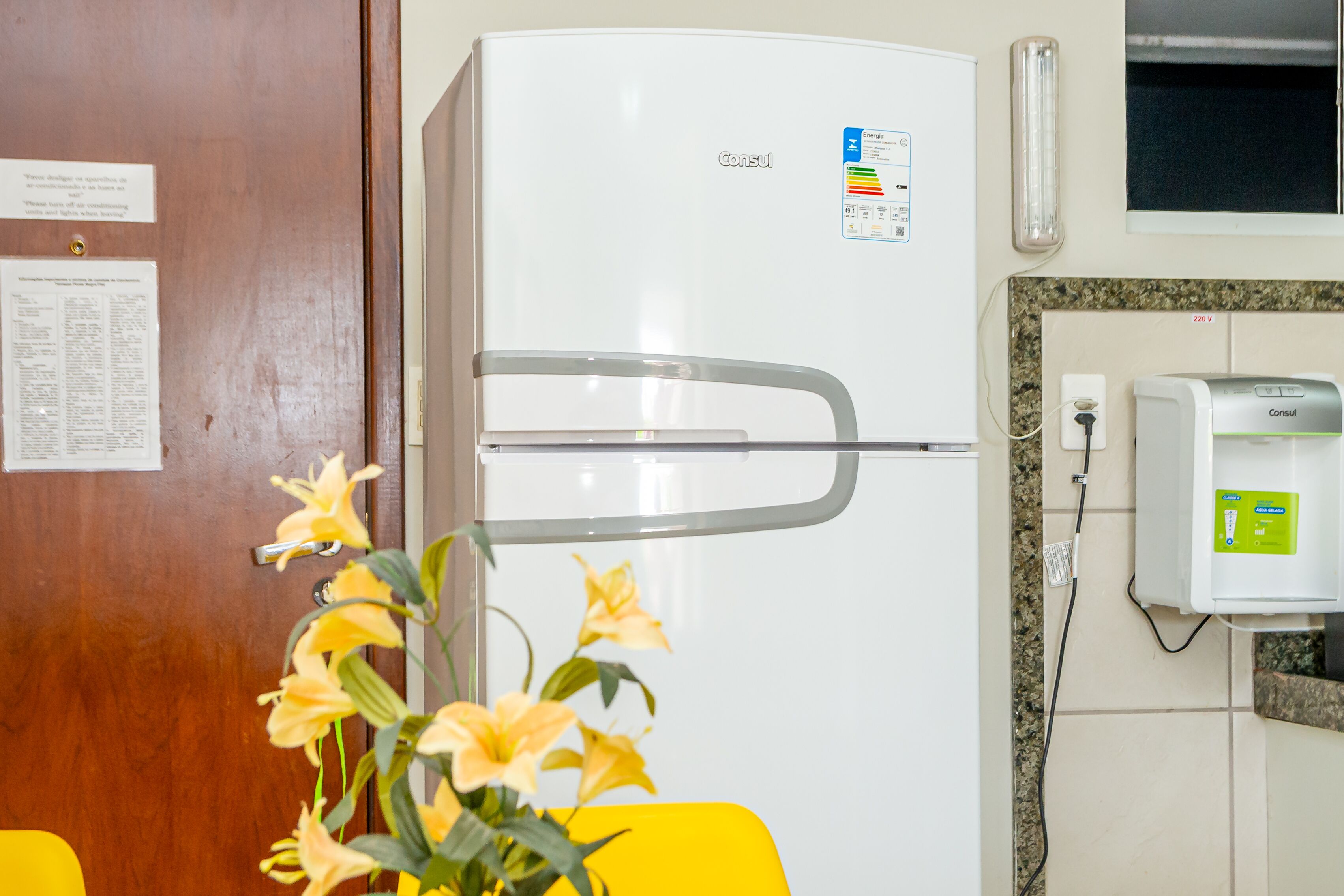 Un réfrigérateur blanc doté d’un distributeur d’eau, une porte en bois avec un avis affiché, et une chaise jaune.