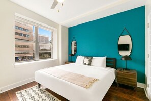 A bedroom with a large bed, a teal accent wall, a window with a view of buildings, and a nightstand with a lamp. - NOLA's Finest: 4-Bedroom Luxury Condo (New Orleans)