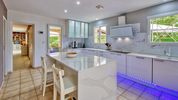 A modern kitchen with a large island, white cabinetry, and a view of greenery outside. - Villa Migron - Charmante maison avec piscine (Migron)