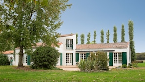 A two-story white building with green shutters and a tiled roof, surrounded by a grassy lawn and trees. - La Maison des Sources - A 20min du Puy du Fou (Mortagne-sur-Sèvre)