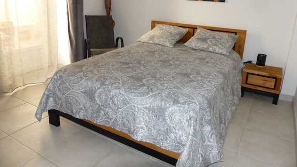A bedroom with a bed, two pillows, a nightstand, a chair, a painting, and a vase.