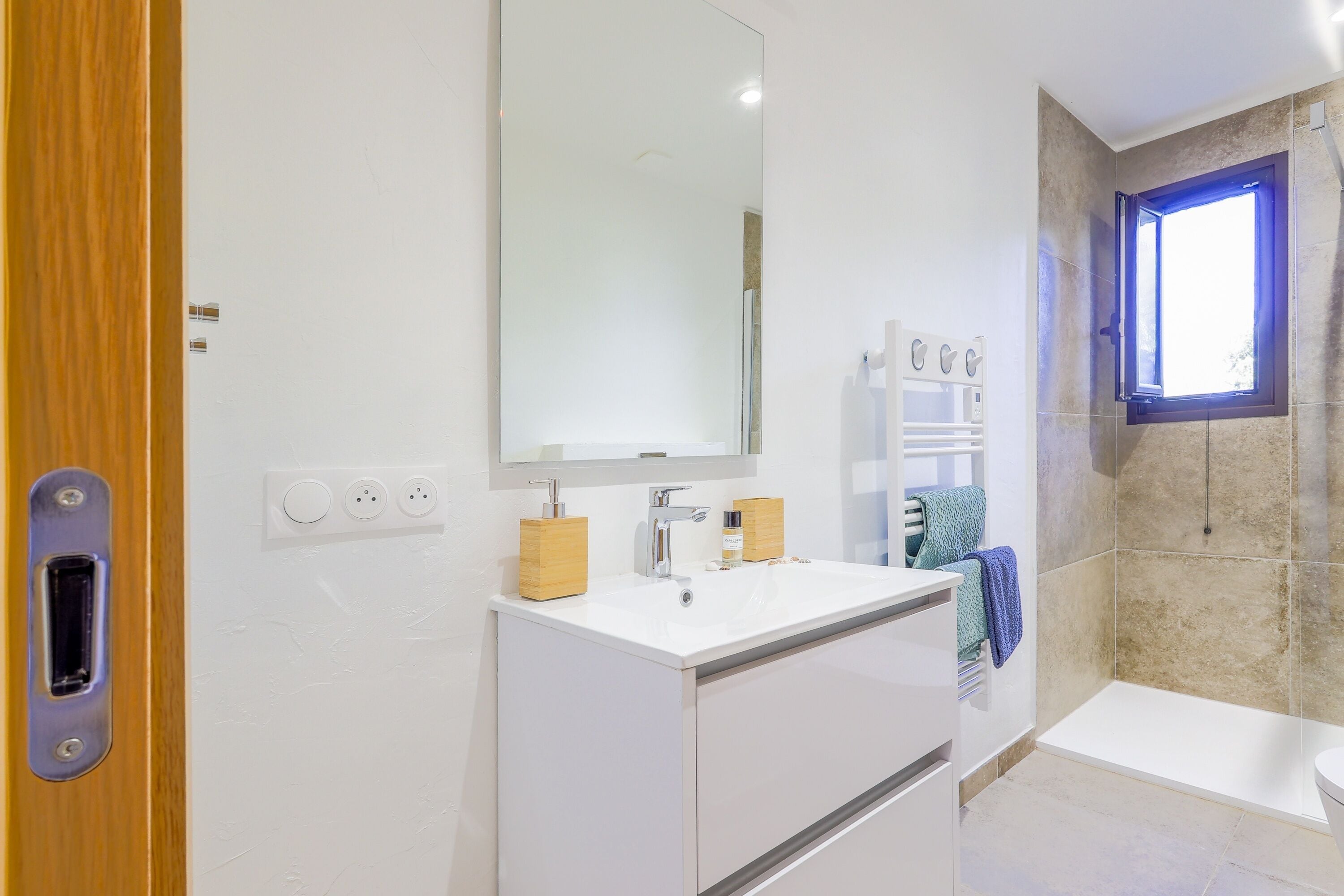 A modern bathroom with a white sink, a mirror, a shower, and a window.
