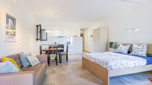 A modern, well-lit room with a bed, a sofa, a dining table with chairs, and a kitchen area in the background. - Petite Pause de Fogata - Jolie vue mer (Corbara)