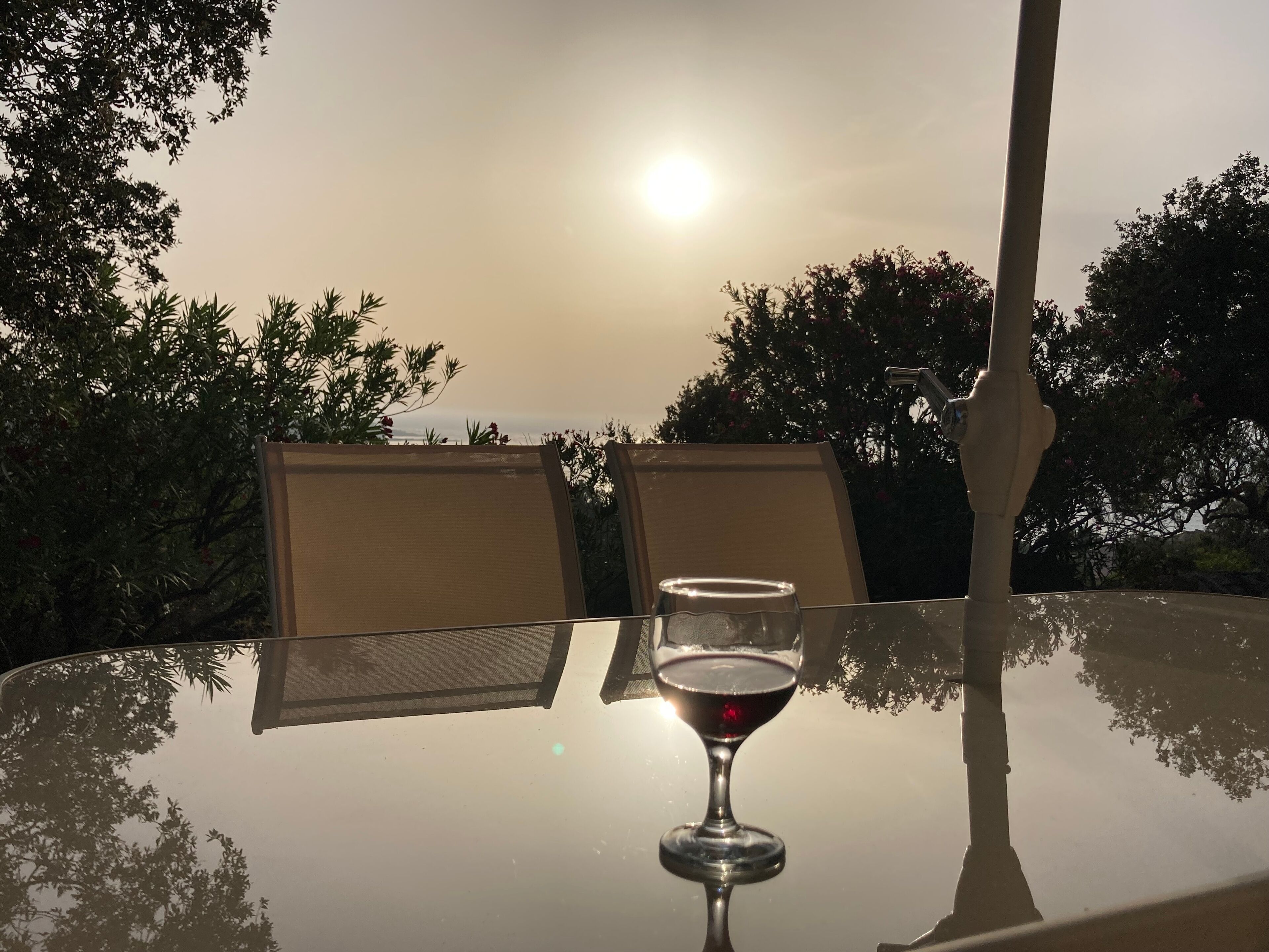 A table with a glass of red wine, two chairs, and a view of the sunset over trees.